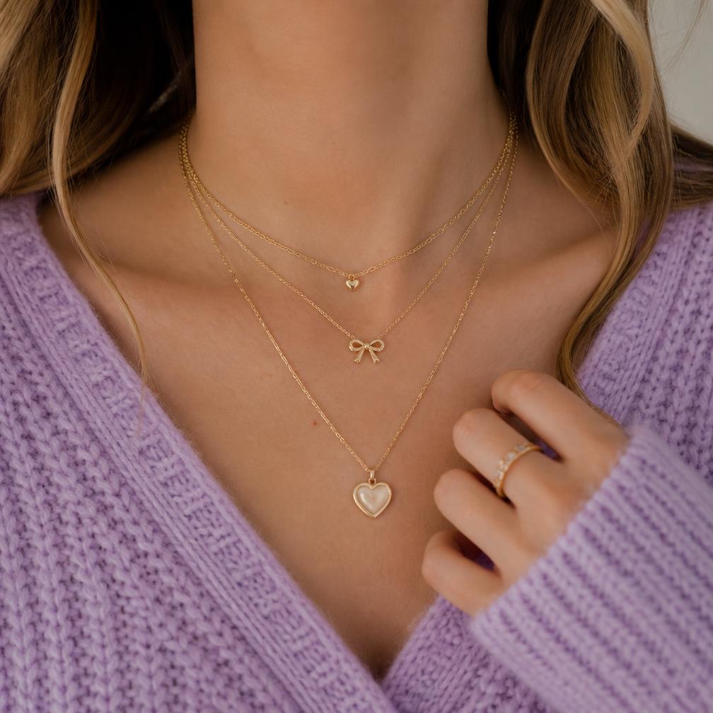 Woman wearing layered dainty gold heart and bow necklaces with a lilac sweater